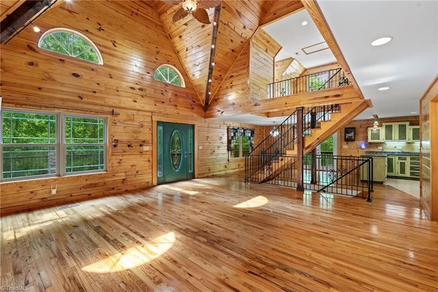 unfurnished living room with light wood-style flooring, stairs, wooden walls, and high vaulted ceiling
