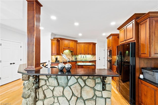 kitchen with ornate columns, black appliances, a kitchen island with sink, crown molding, and light wood-type flooring