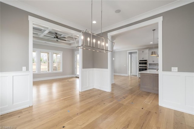 unfurnished dining area with a wainscoted wall, coffered ceiling, light wood-style flooring, beamed ceiling, and ceiling fan with notable chandelier