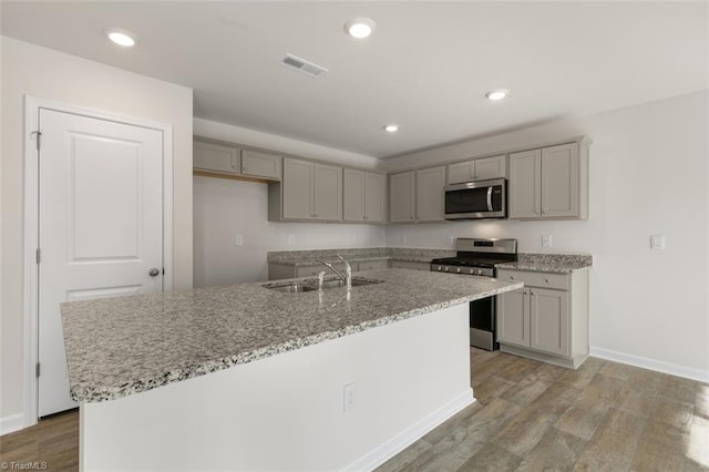 kitchen featuring appliances with stainless steel finishes, light wood-type flooring, light stone counters, sink, and an island with sink