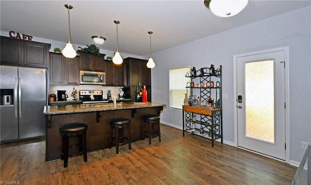 kitchen featuring stainless steel appliances, an island with sink, dark brown cabinets, dark stone counters, and a breakfast bar