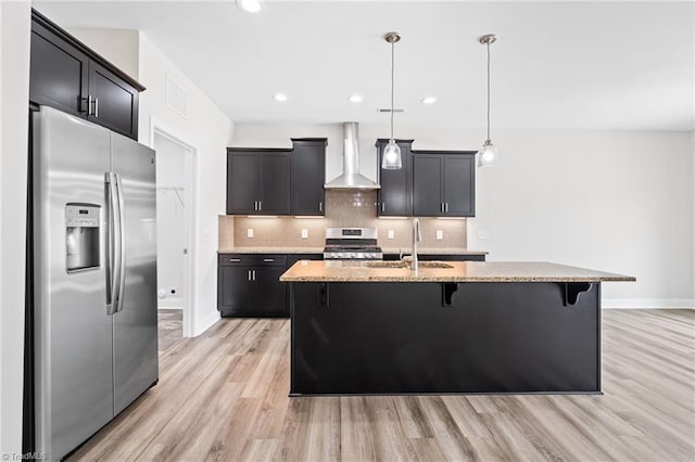 kitchen with stainless steel appliances, light stone counters, wall chimney exhaust hood, and an island with sink