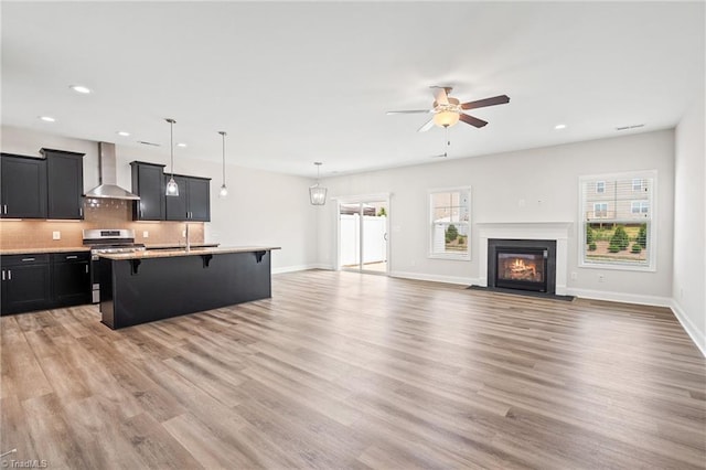 kitchen with a breakfast bar, a kitchen island with sink, hanging light fixtures, wall chimney exhaust hood, and stainless steel range oven