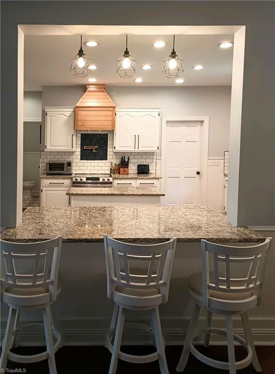 kitchen featuring custom range hood, white cabinets, decorative backsplash, decorative light fixtures, and a kitchen bar