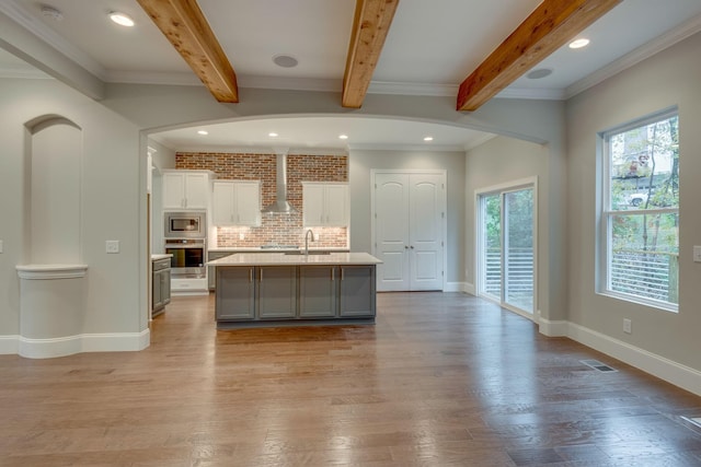 kitchen featuring crown molding, light hardwood / wood-style flooring, a center island with sink, wall chimney exhaust hood, and tasteful backsplash