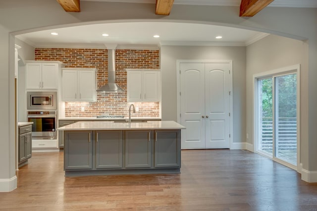 kitchen featuring light hardwood / wood-style floors, stainless steel appliances, a center island with sink, wall chimney range hood, and white cabinetry