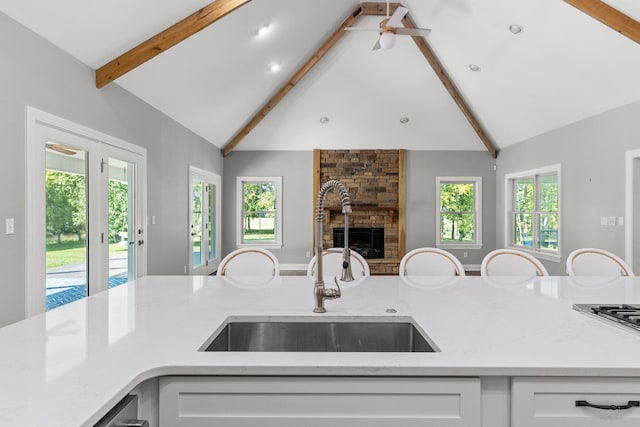 kitchen featuring beam ceiling, a stone fireplace, light stone counters, white cabinetry, and a sink