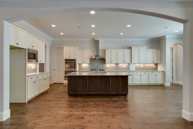 kitchen featuring backsplash, appliances with stainless steel finishes, an island with sink, wall chimney range hood, and dark hardwood / wood-style flooring