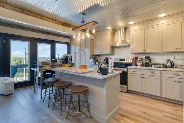 kitchen with stainless steel electric range oven, wall chimney exhaust hood, light hardwood / wood-style flooring, an island with sink, and decorative light fixtures