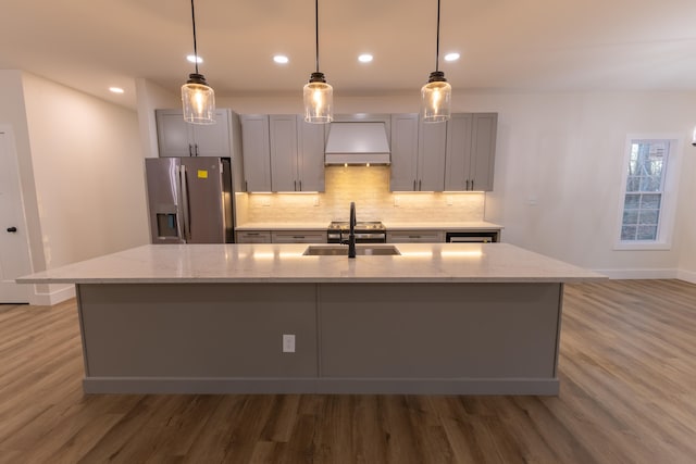 kitchen with an island with sink, premium range hood, light stone counters, and stainless steel fridge with ice dispenser