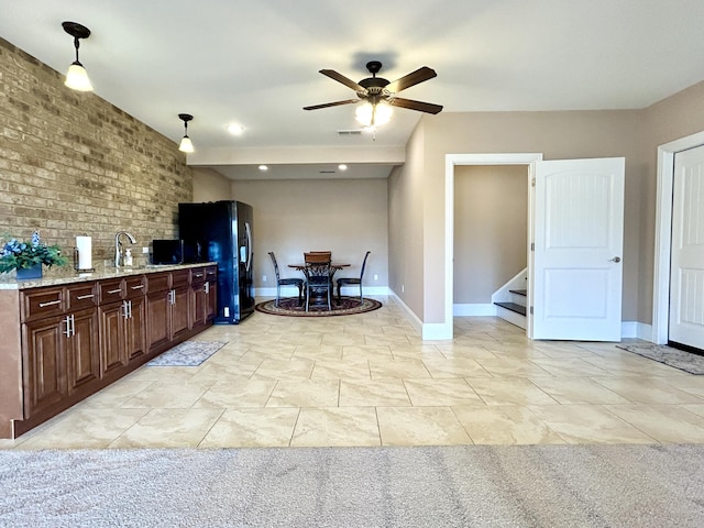 kitchen with ceiling fan, hanging light fixtures, light stone countertops, brick wall, and black refrigerator with ice dispenser