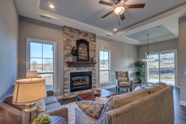 living room featuring a stone fireplace, a raised ceiling, dark wood-type flooring, and ceiling fan with notable chandelier