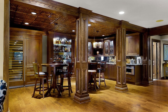 bar with light wood-type flooring, an ornate ceiling, wet bar, and ornate columns