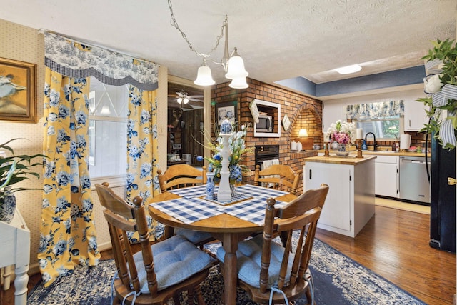 dining area with sink, ceiling fan, a brick fireplace, dark wood-type flooring, and a textured ceiling