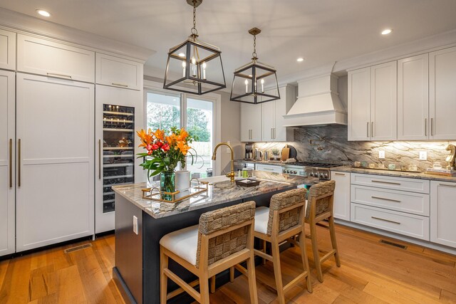 kitchen with a breakfast bar, light stone counters, a kitchen island with sink, light wood-type flooring, and custom range hood