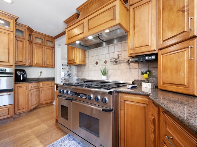 kitchen with custom range hood, dark stone countertops, light wood-type flooring, decorative backsplash, and appliances with stainless steel finishes