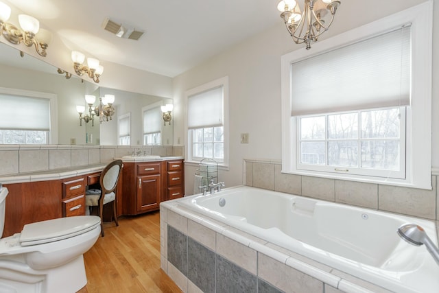 bathroom featuring toilet, wood-type flooring, vanity, a notable chandelier, and a relaxing tiled tub