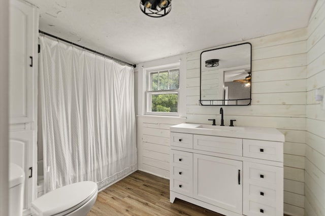 bathroom with wood-type flooring, wooden walls, vanity, ceiling fan, and toilet