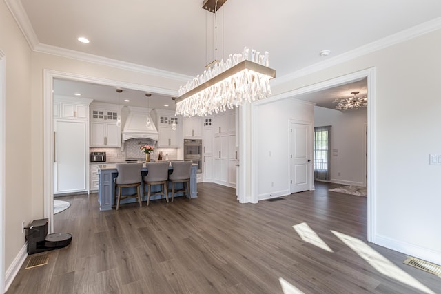 kitchen featuring custom range hood, hanging light fixtures, glass insert cabinets, white cabinetry, and a kitchen island with sink