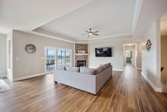 living room with ceiling fan, a tray ceiling, hardwood / wood-style floors, and a brick fireplace