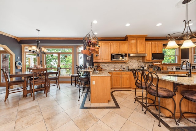 kitchen with light stone countertops, a notable chandelier, a kitchen island with sink, hanging light fixtures, and stainless steel microwave