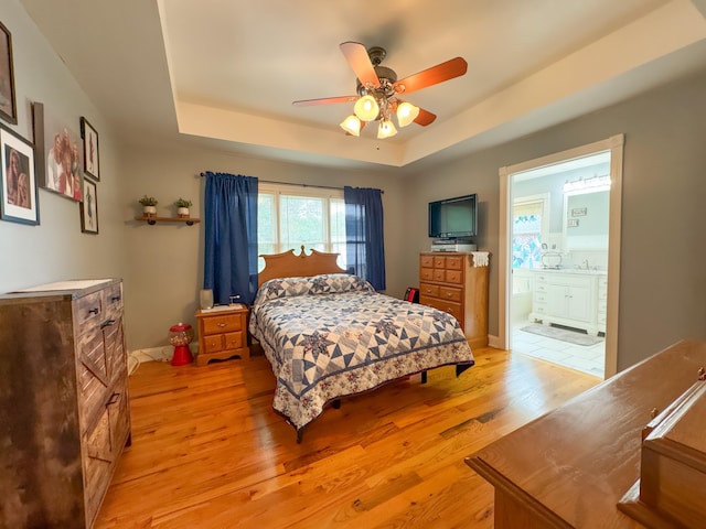 bedroom with a raised ceiling, ceiling fan, connected bathroom, and light hardwood / wood-style floors