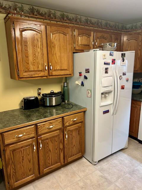kitchen featuring white refrigerator with ice dispenser, dark countertops, brown cabinetry, and light tile patterned floors