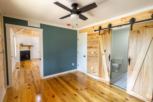 unfurnished bedroom featuring ceiling fan, a barn door, ensuite bathroom, wood walls, and hardwood / wood-style flooring