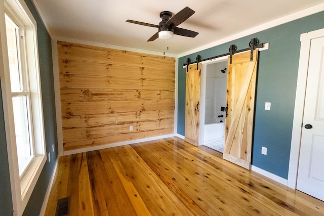 unfurnished bedroom with ceiling fan, a barn door, wood walls, hardwood / wood-style flooring, and ornamental molding