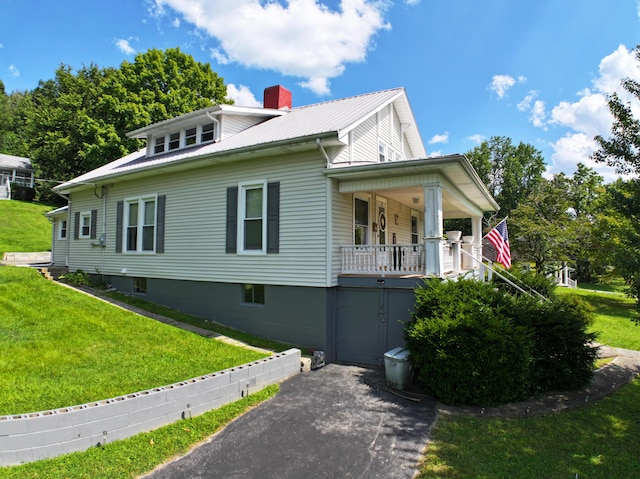 view of front of property featuring metal roof, aphalt driveway, a front yard, covered porch, and a chimney