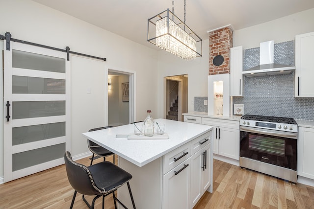 kitchen with light wood-type flooring, a barn door, a breakfast bar area, wall chimney range hood, and stainless steel range with gas stovetop