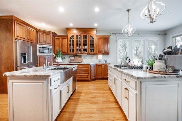 kitchen with an island with sink, pendant lighting, stainless steel appliances, light wood-type flooring, and an inviting chandelier