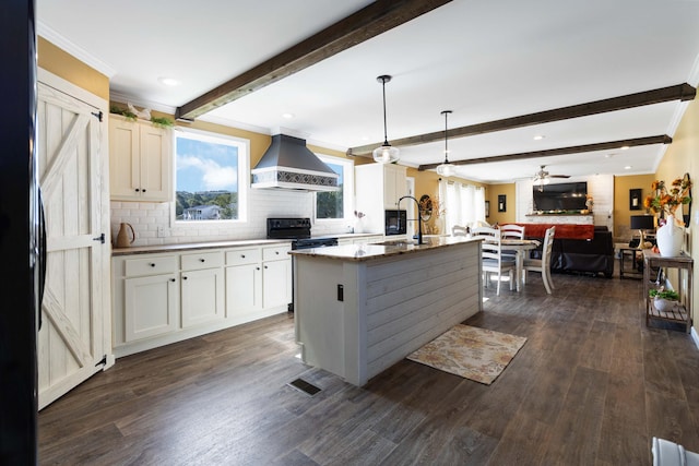 kitchen with backsplash, an island with sink, ceiling fan, dark wood-type flooring, and wall chimney exhaust hood