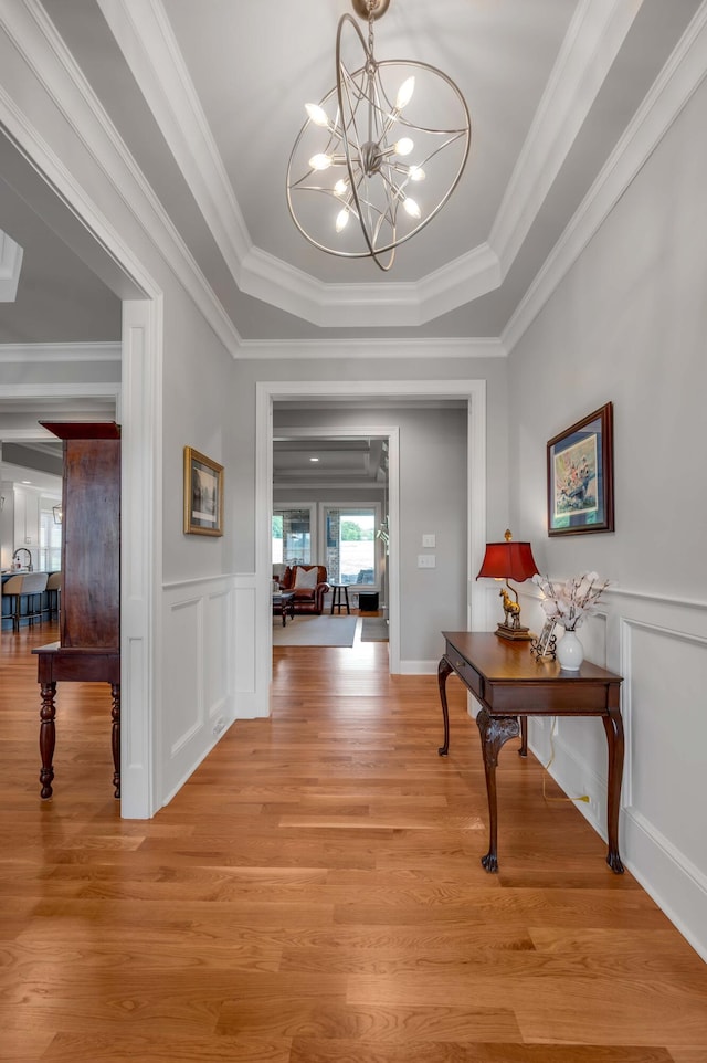 hallway featuring a notable chandelier, ornamental molding, a tray ceiling, light wood-style floors, and a decorative wall