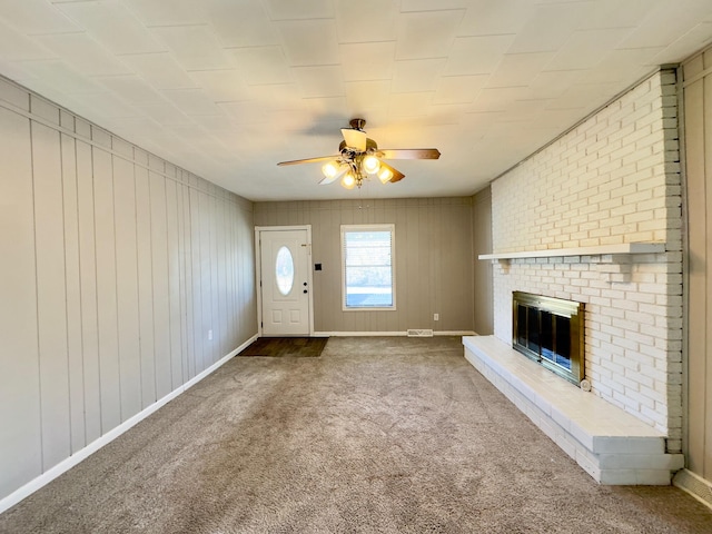 unfurnished living room featuring wooden walls, a brick fireplace, ceiling fan, and carpet flooring