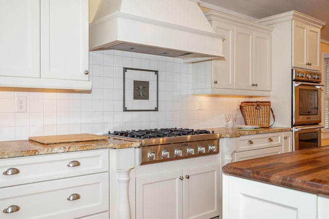 kitchen with white cabinets, appliances with stainless steel finishes, custom exhaust hood, backsplash, and light stone counters