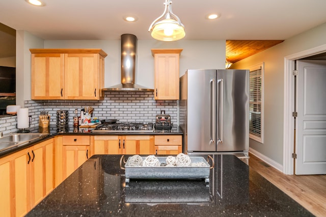 kitchen featuring light brown cabinetry, dark stone countertops, backsplash, stainless steel appliances, and wall chimney exhaust hood