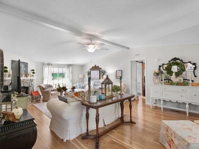 living room with ceiling fan, a stone fireplace, a textured ceiling, lofted ceiling with beams, and light wood-type flooring