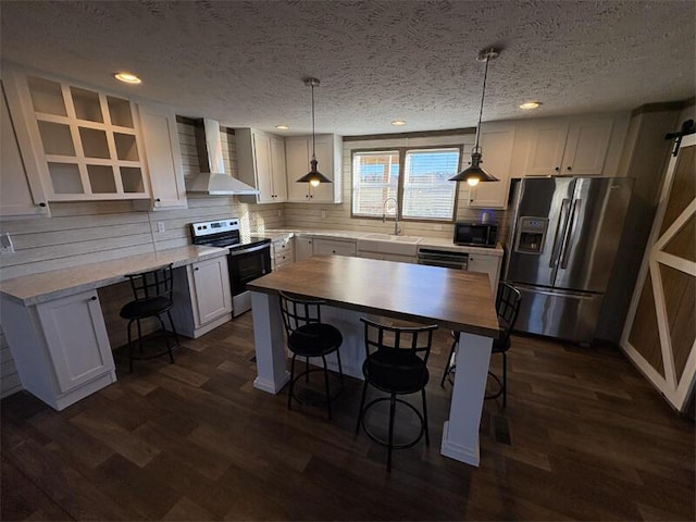 kitchen with a textured ceiling, stainless steel appliances, wall chimney range hood, pendant lighting, and white cabinetry