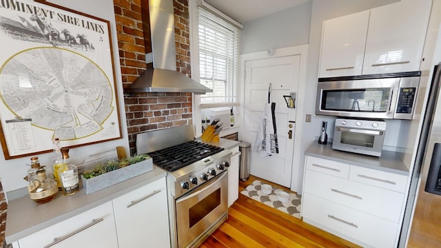 kitchen with white cabinets, brick wall, light hardwood / wood-style flooring, wall chimney range hood, and appliances with stainless steel finishes