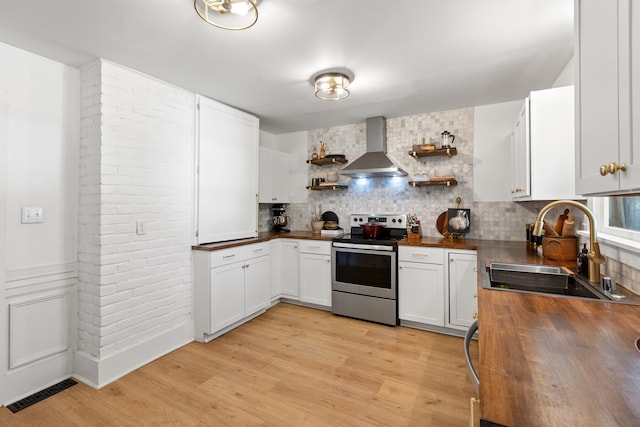 kitchen featuring stainless steel range with electric stovetop, wall chimney exhaust hood, sink, butcher block countertops, and white cabinetry