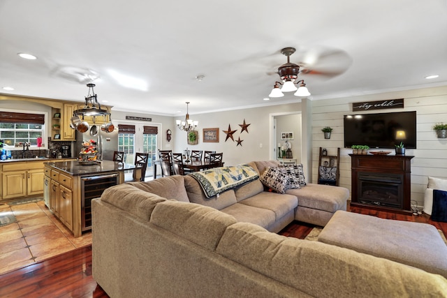 living room with ceiling fan with notable chandelier, crown molding, sink, wood-type flooring, and beverage cooler