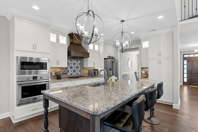 kitchen with a kitchen island with sink, sink, appliances with stainless steel finishes, premium range hood, and white cabinetry