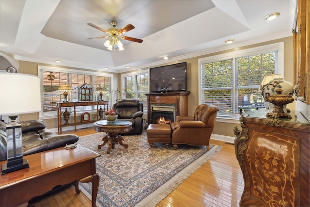 living room featuring a warm lit fireplace, light wood-type flooring, a raised ceiling, and a ceiling fan