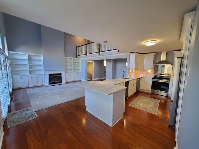 kitchen featuring wall chimney range hood, decorative light fixtures, kitchen peninsula, appliances with stainless steel finishes, and white cabinetry