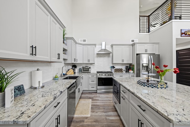 kitchen with light stone countertops, stainless steel appliances, wall chimney exhaust hood, a towering ceiling, and light hardwood / wood-style flooring