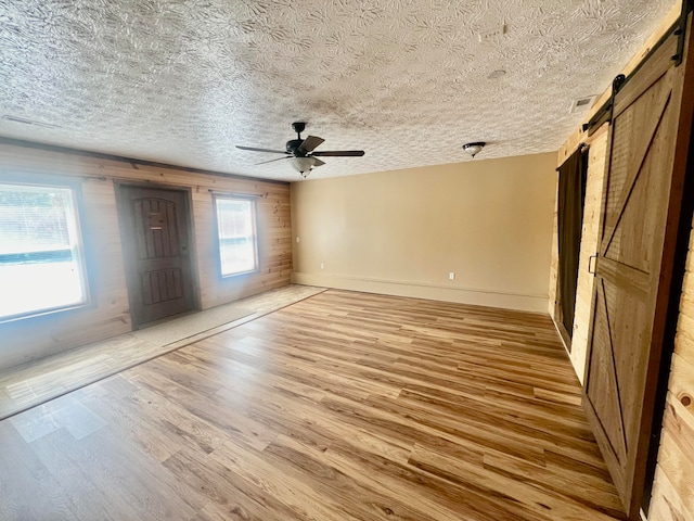 unfurnished bedroom featuring ceiling fan, light hardwood / wood-style floors, a barn door, wood walls, and a textured ceiling