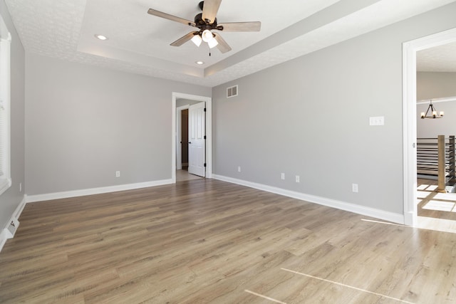 spare room featuring ceiling fan with notable chandelier, a tray ceiling, visible vents, wood finished floors, and baseboards