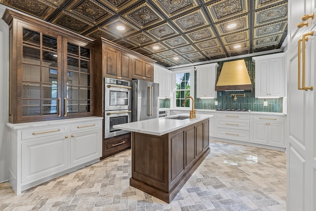 kitchen featuring a kitchen island with sink, stainless steel appliances, white cabinets, custom range hood, and sink