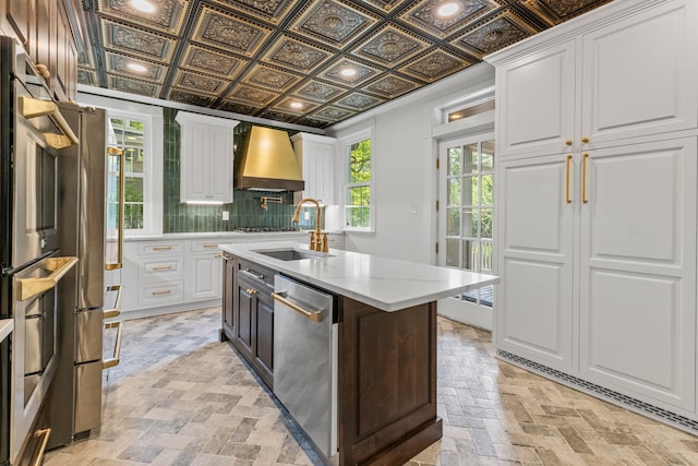 kitchen with an island with sink, sink, white cabinetry, custom exhaust hood, and appliances with stainless steel finishes